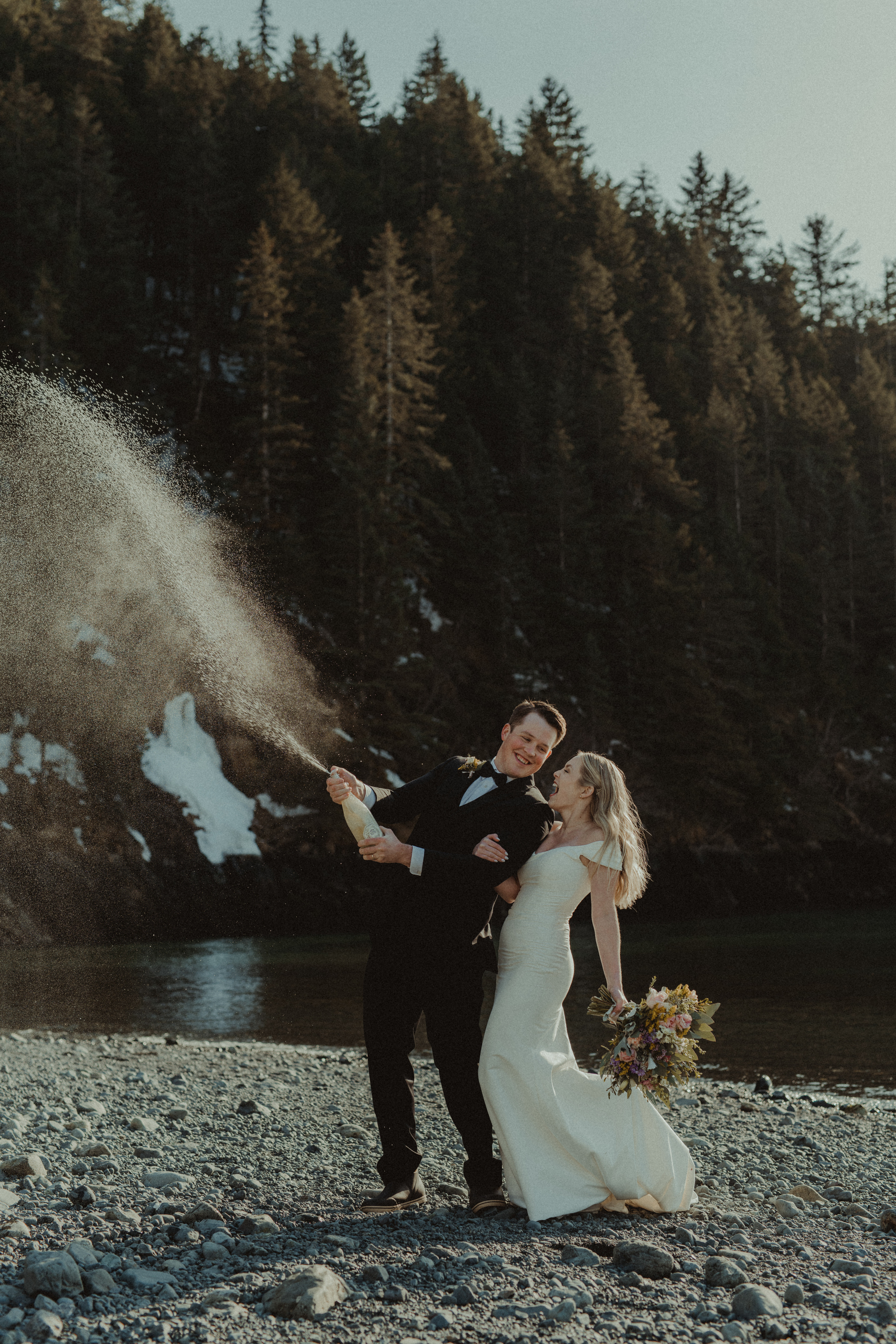 A bride and groom pop a bottle of champagne on a remote beach in Alaska on the coast of prince william sound