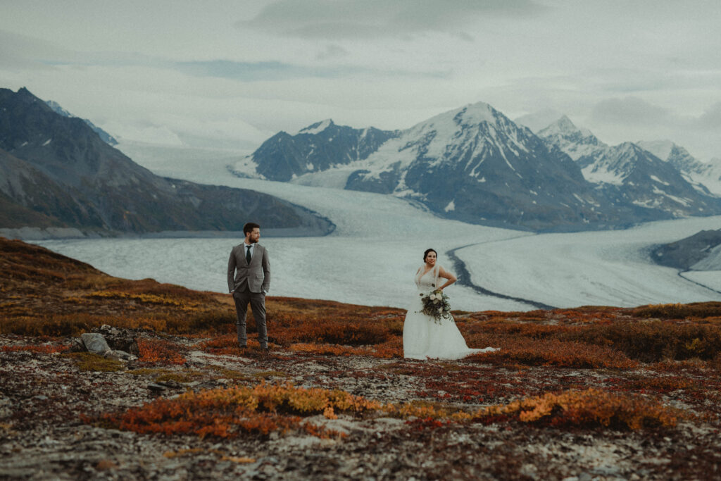 Couple stand on a mountaintop with red and orange fall foliage around them. Colony Glacier rests in the background. 