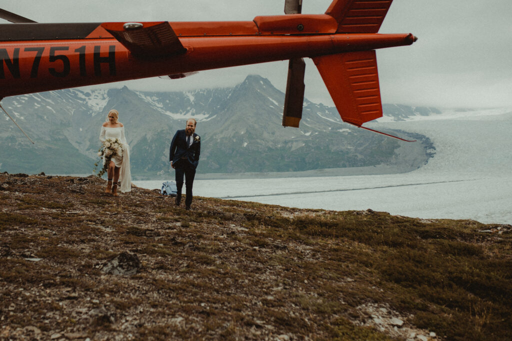 Bride and groom stand in the distance on a mountain top with a helicopter in the forefront
