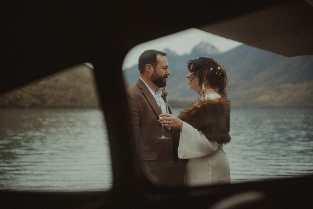 Bride and groom share a smile and glass of champagne against the backdrop of Eklutna lake